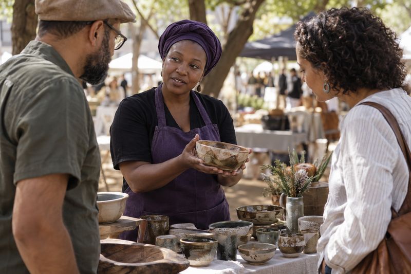 AI-generated concept: A ceramic maker in an apron shows a bowl to two visitors at her market stall.